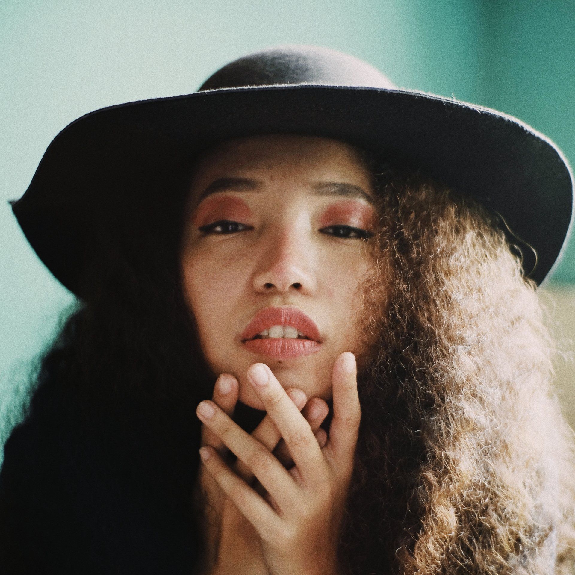 Woman with curly hair, dark hat, wearing red eyeshadow, and holding hands near her face.