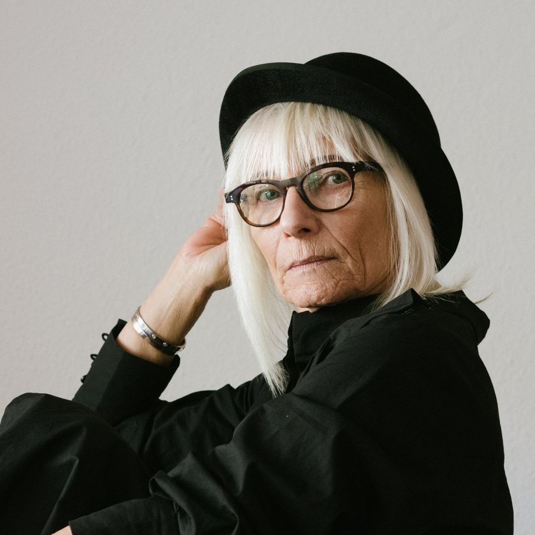Woman with white hair, black hat and glasses, looking at the camera.