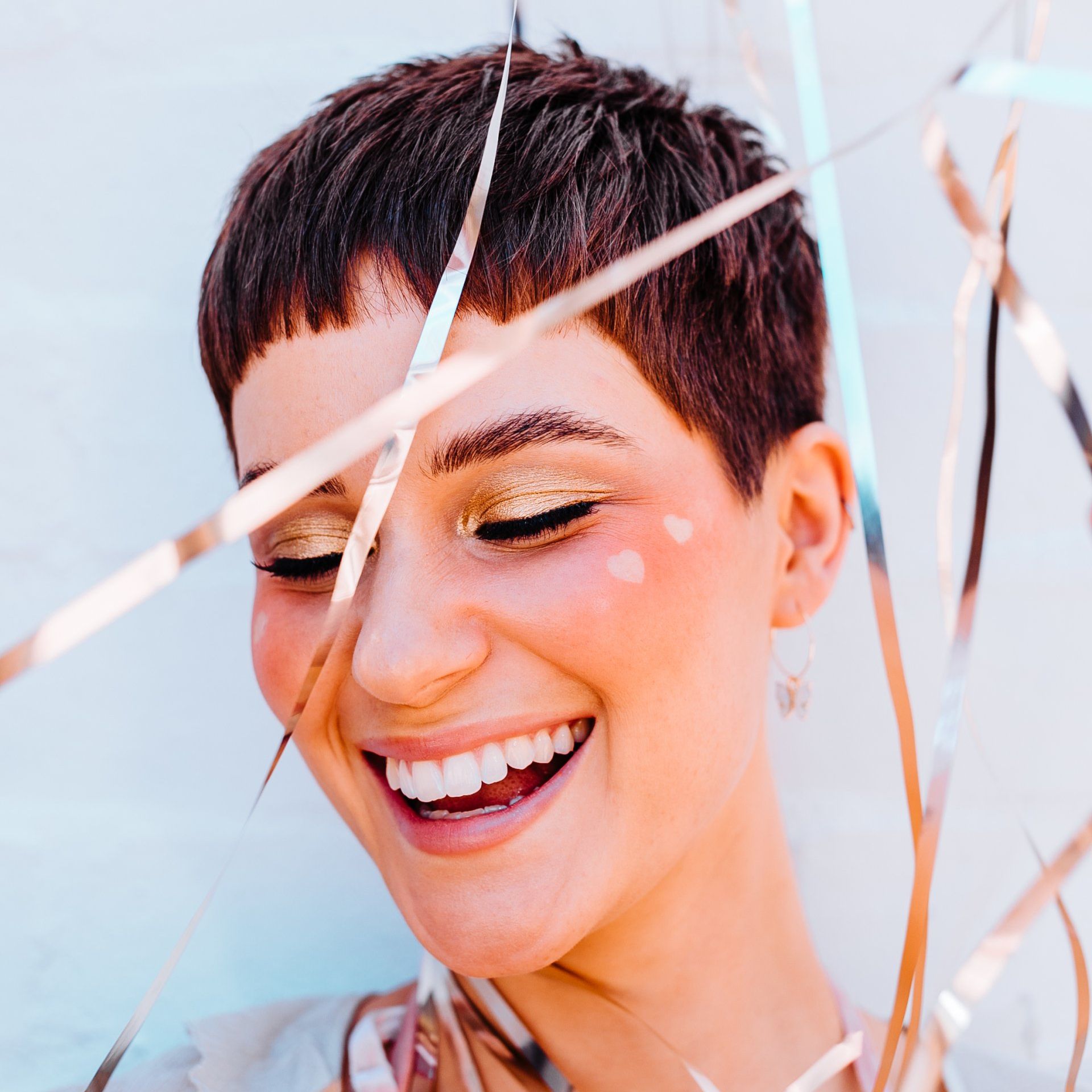 Woman with short brown hair, smiling with golden eyeshadow, pink cheek accents, and party streamers.