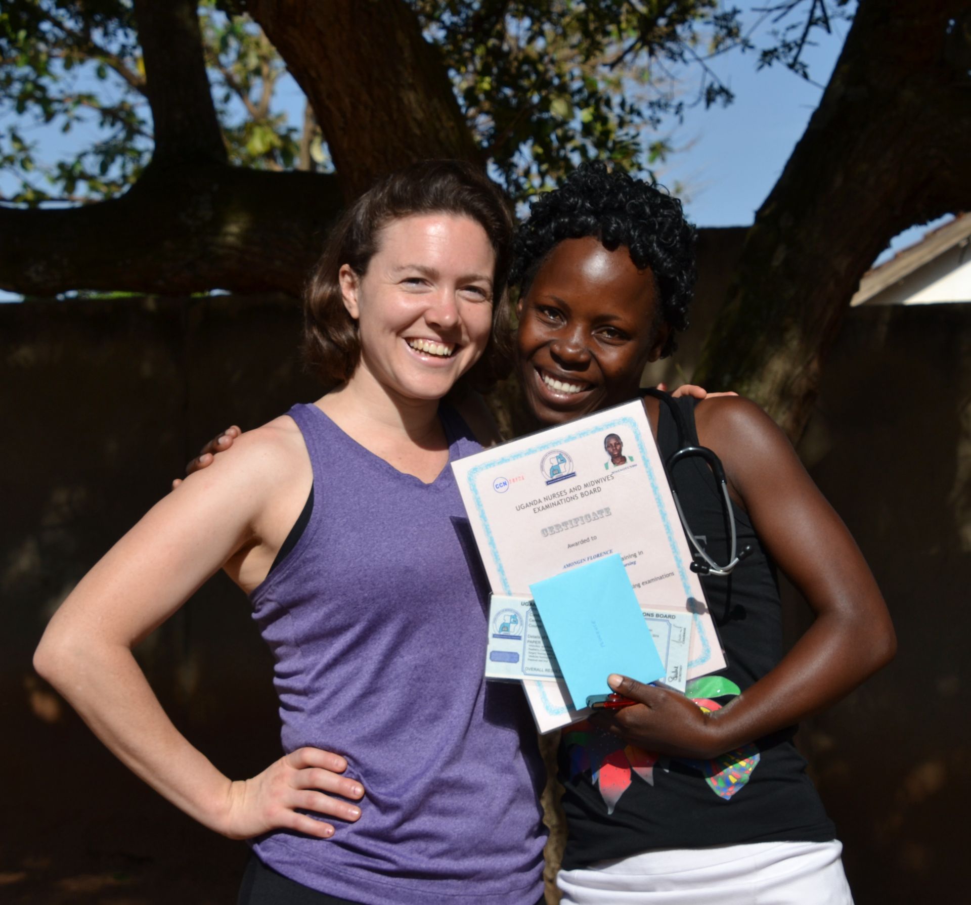 Two women smiling, one holding a certificate and blue booklet, in front of a tree.