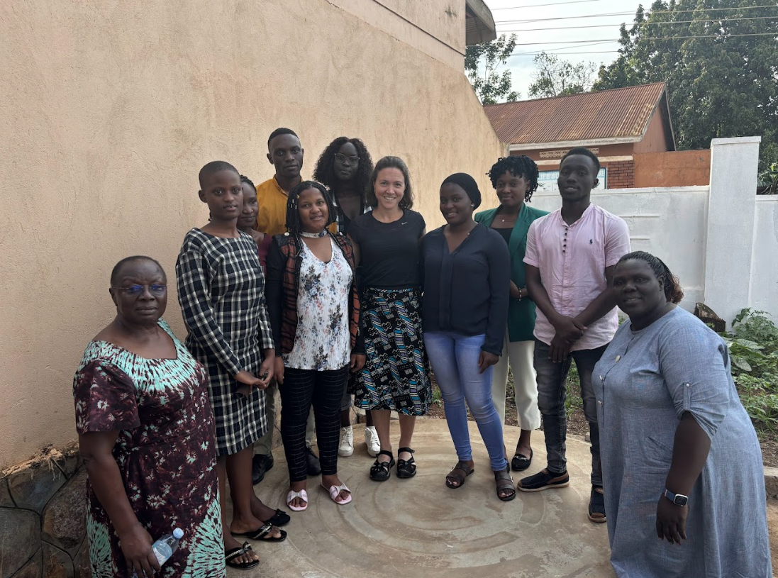 Group of people standing in front of a building. Some are smiling, and the colors are neutral with green vegetation.