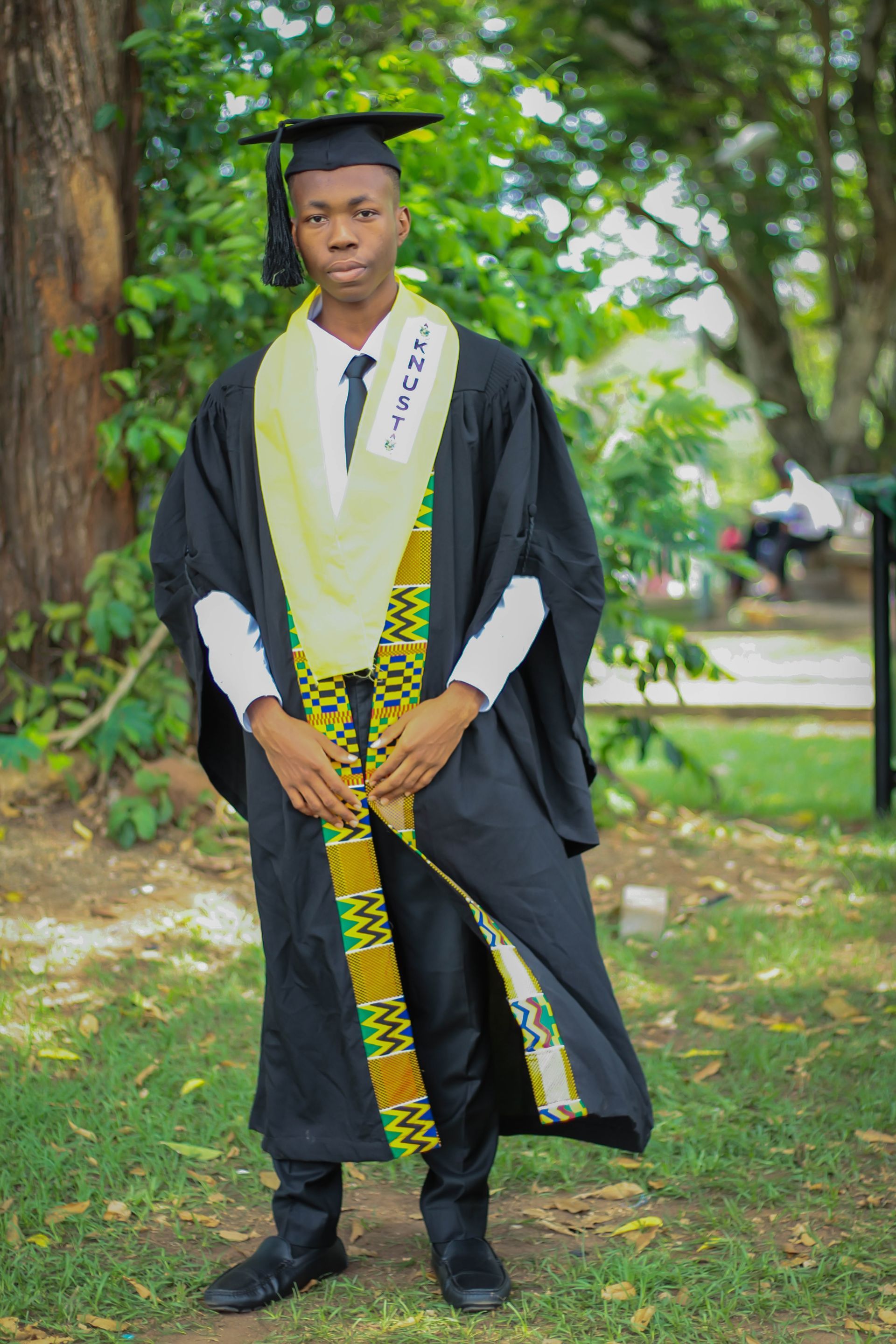 Person in graduation gown stands outdoors on grass, wearing a yellow sash with black pattern.