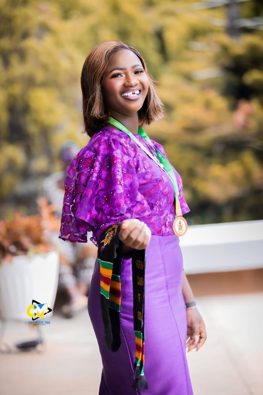Woman in purple outfit smiling, holding sash, wearing medals. Outdoor setting.