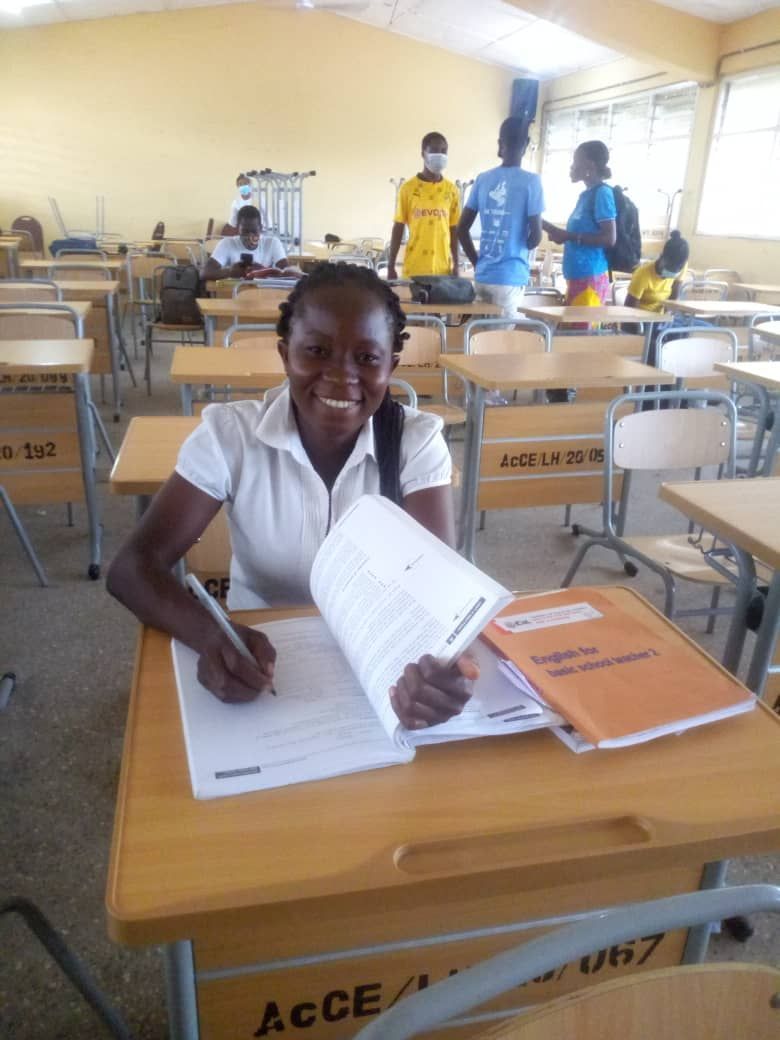 Student writing at desk in a classroom, smiling. Others stand in the background. Orange book visible.