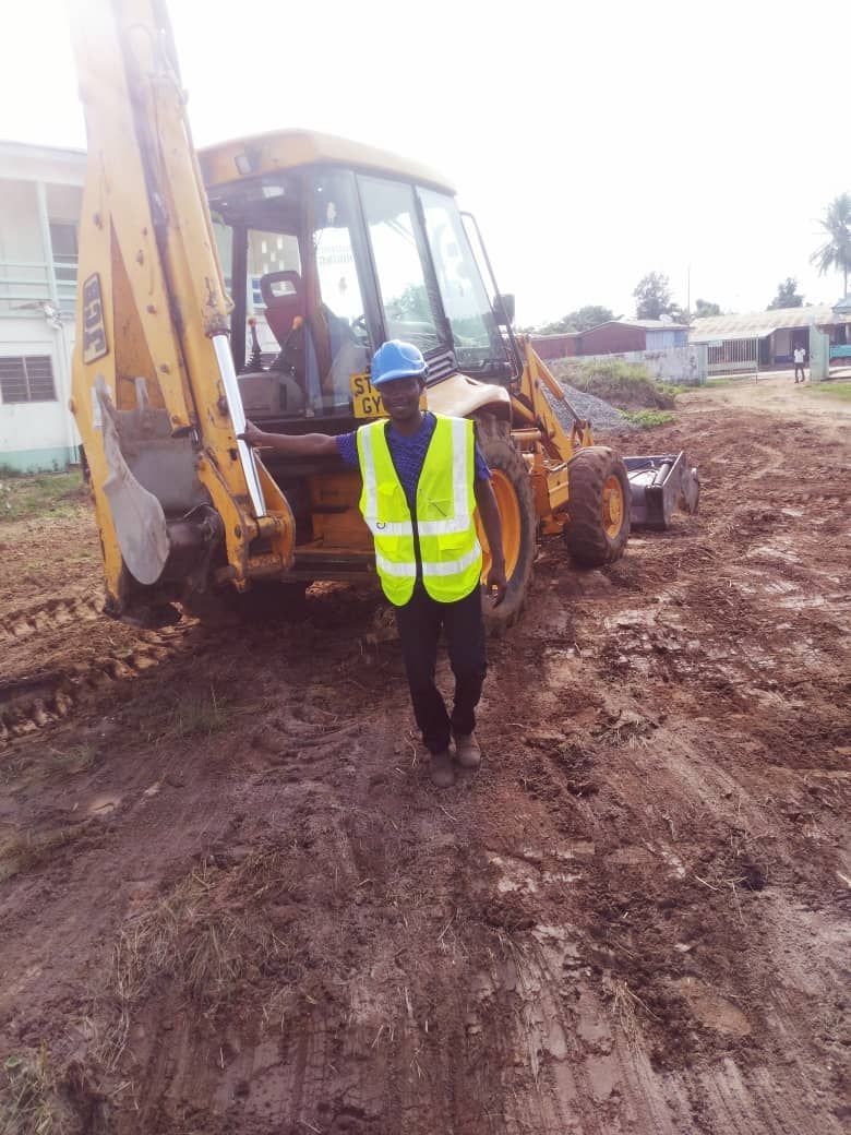 Man in safety vest stands in front of a yellow backhoe on muddy ground. Another person is in the cab.