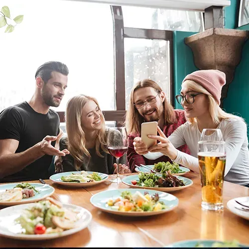 Four people at a table in a restaurant. One shows phone to the group. Food and drinks are on the table.