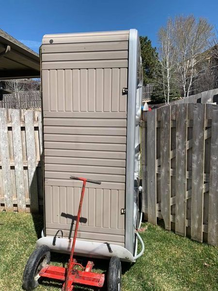 A portable toilet is being pushed by a red wagon in a backyard.