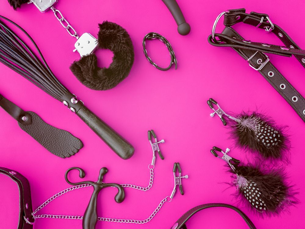 An assortment of black BDSM-related items, including cuffs, a whip, and clamps, arranged on a vibrant pink background — A Bit Cheeky The Adult Store In Lismore, NSW