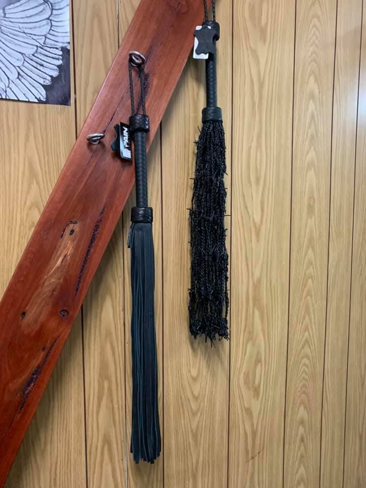 Two Black Whips Hang Against a Wood-paneled Wall — A Bit Cheeky The Adult Store In Lismore, NSW