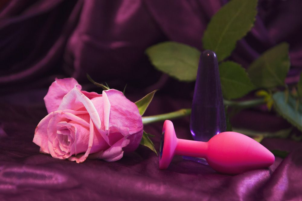 A pink rose, a translucent purple tapered item, and a bright pink plug-shaped object on a purple satin fabric background — A Bit Cheeky The Adult Store In Lismore, NSW