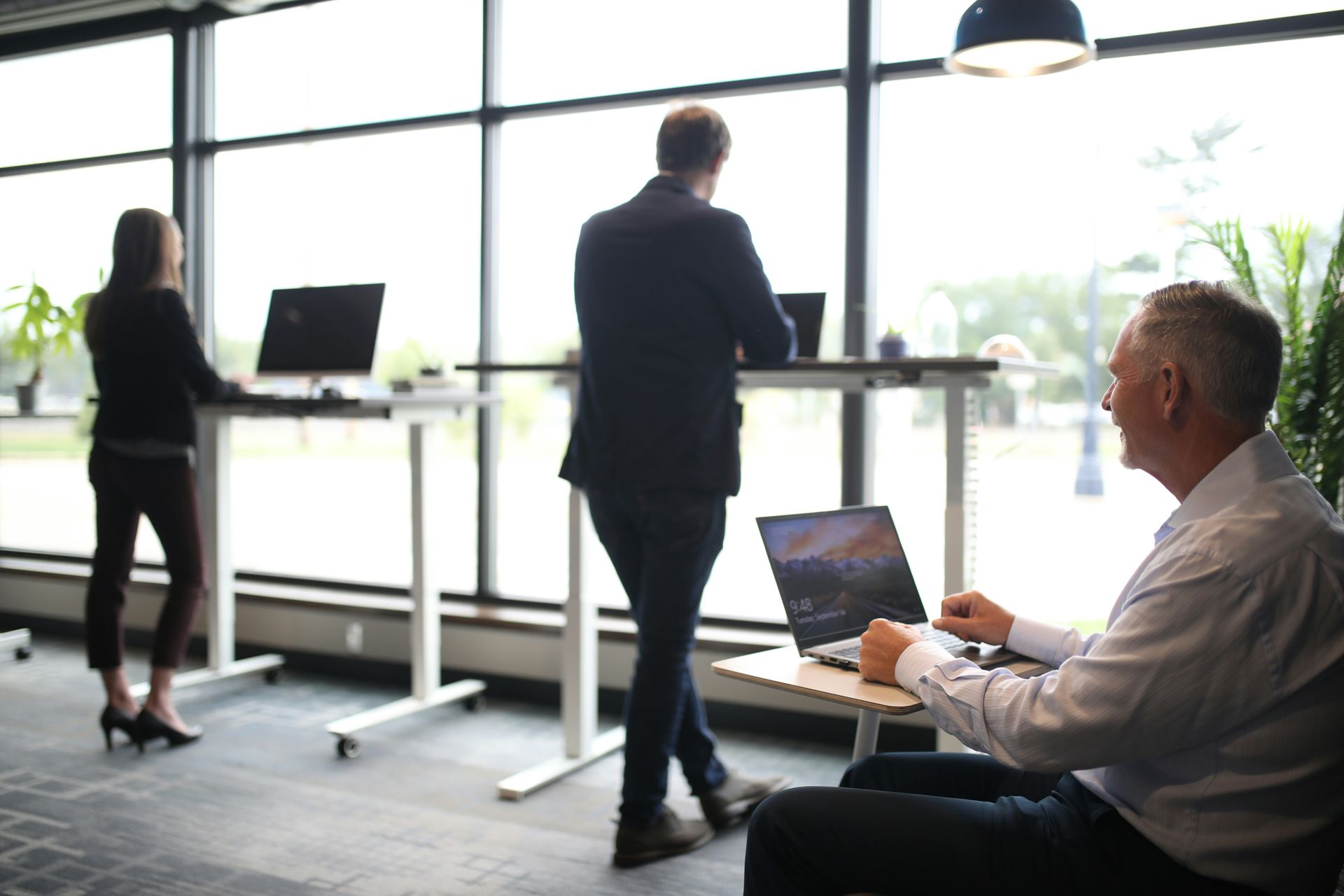 Three people working in modern office by large window; two stand at desks, one seated with laptop.