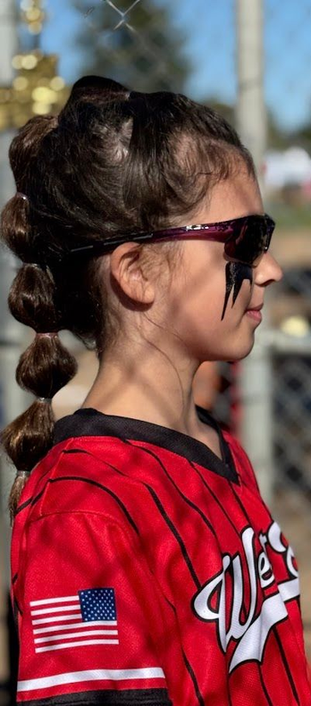 A person wearing a hat and orange shirt, holding a baseball bat on their shoulder on a baseball field.