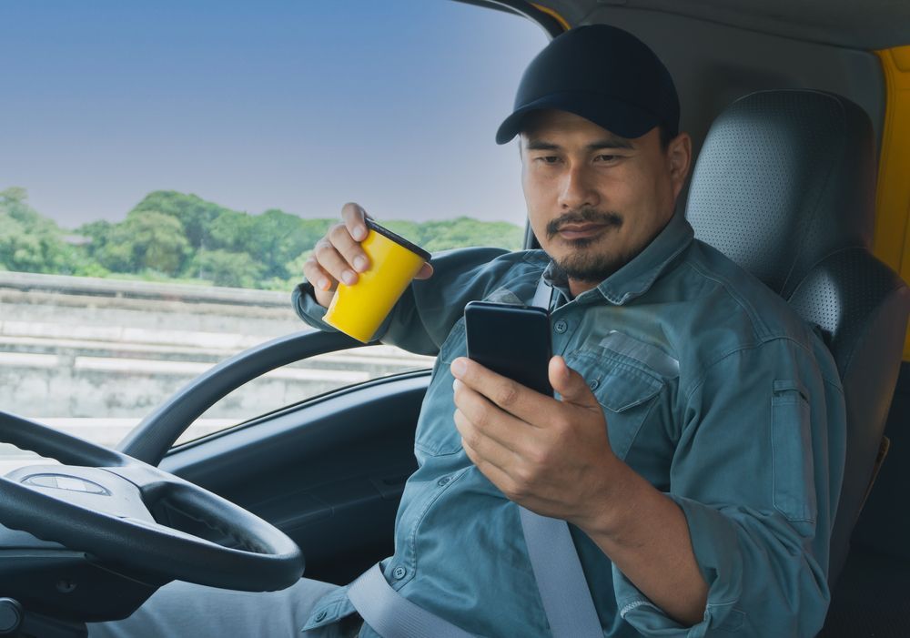 a man is sitting in the driver 's seat of a truck holding a cup of coffee and looking at his cell phone .