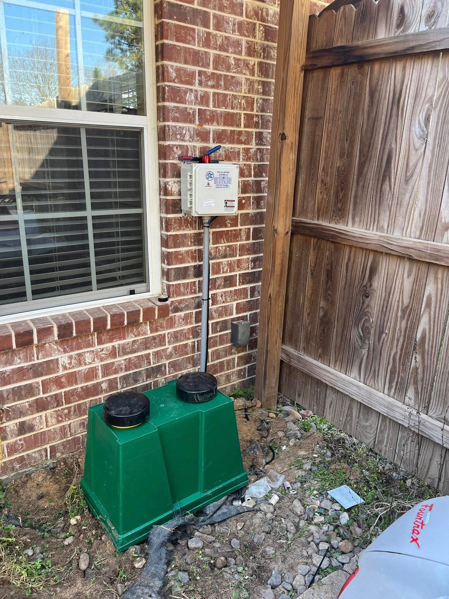 A green box is sitting in front of a brick building next to a wooden fence.