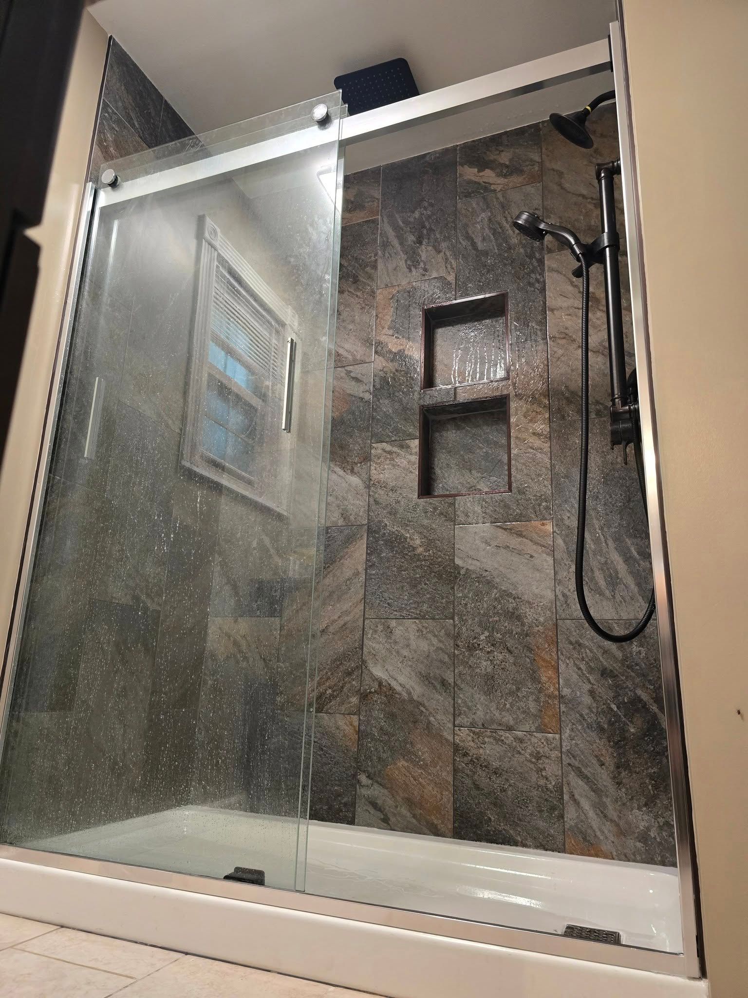 Glass shower stall with stone-look tile walls and a black handheld shower head