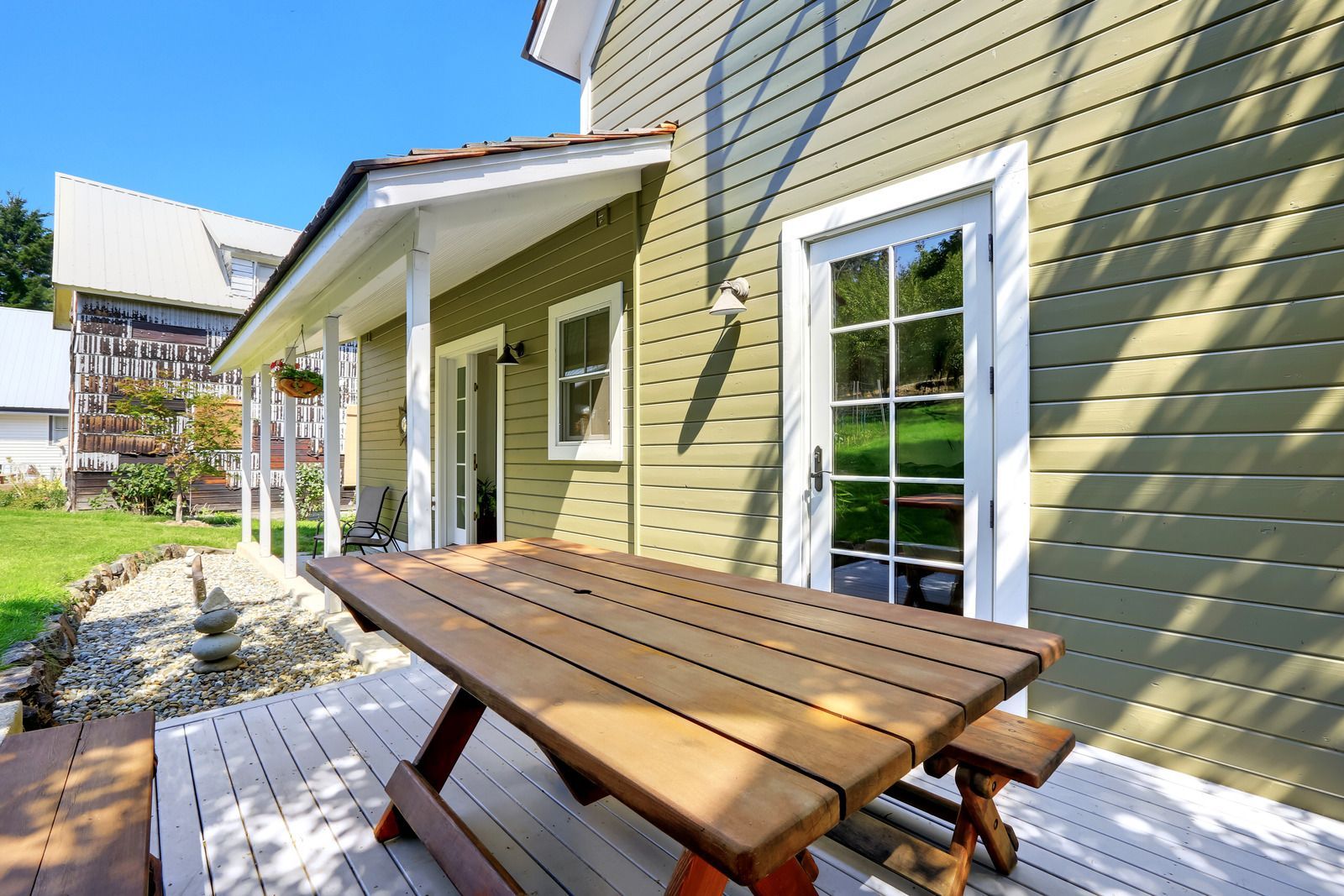 Wooden picnic table on a patio beside a green house with white-trim windows.