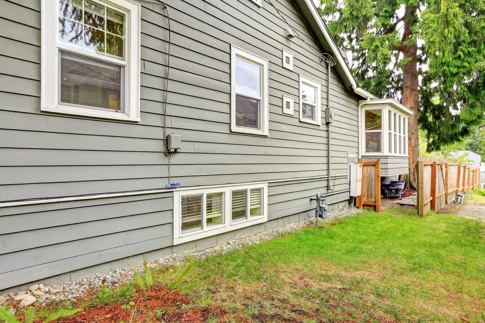 Side view of a gray house with windows, air-conditioning units, and a small fenced yard with grass and trees.