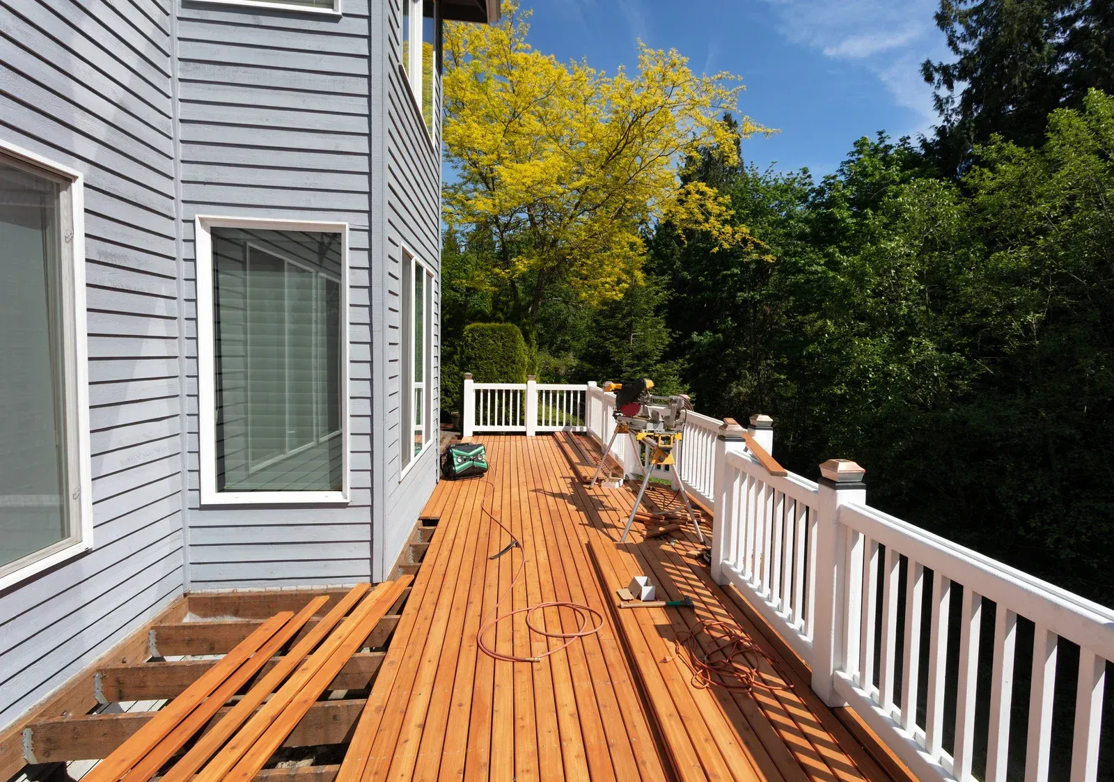 Wooden deck beside a gray house, with white railing, green trees, and blue sky.