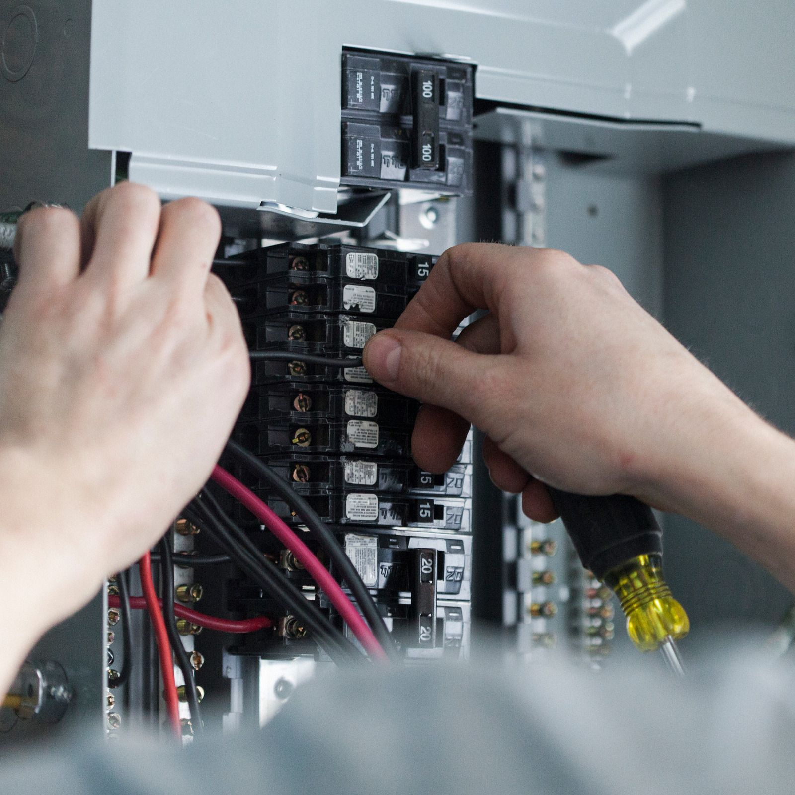 Hands wiring an open electrical breaker panel with red and black cables and a screwdriver