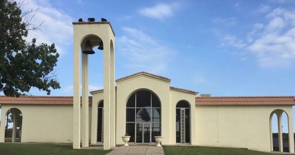 Off-white building with bell tower, arched doorways, glass front, and red-tiled roof under a blue sky.