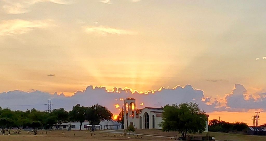 Sunset over a light-colored church with a bell tower. Trees and a cemetery in the foreground; orange and yellow sky.