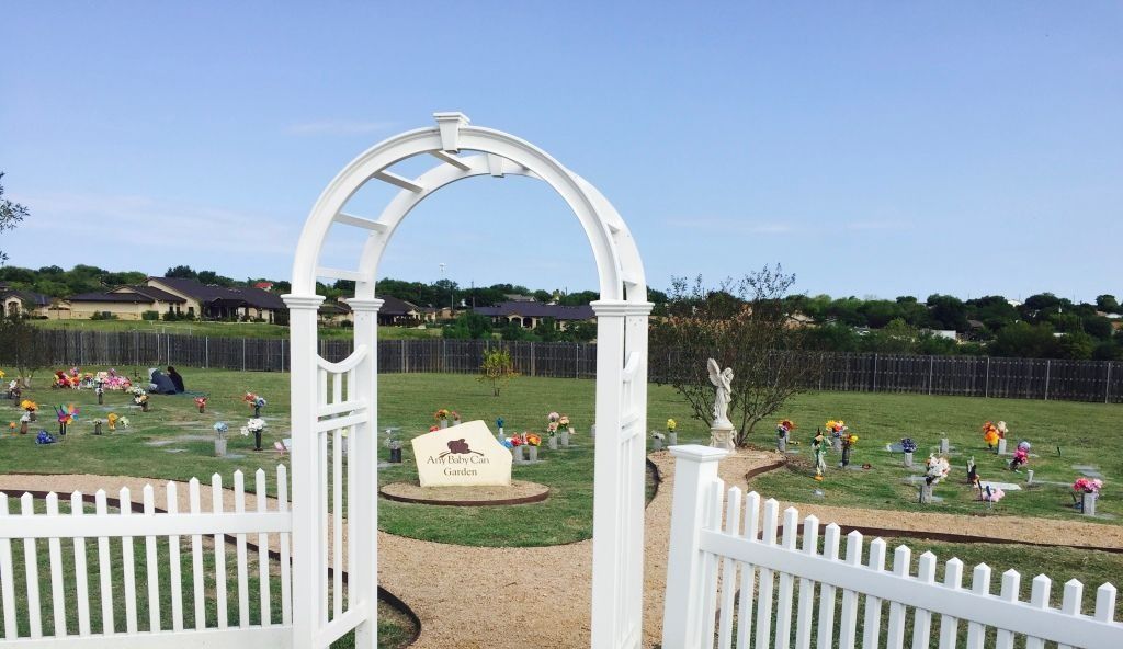 White archway entrance to a cemetery. Headstones, grass, and a few houses in the background.