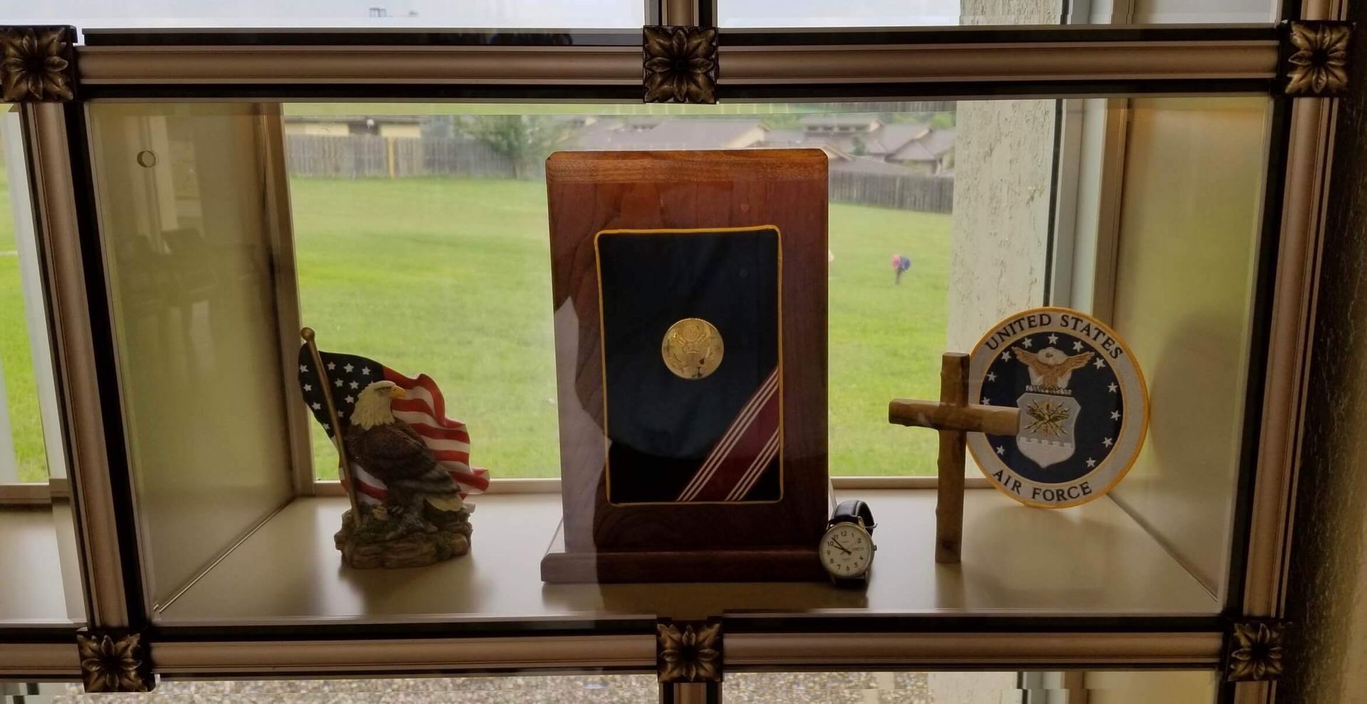 Display case with U.S. flag, bald eagle statue, military insignia, cross, and framed medal, in front of a window.
