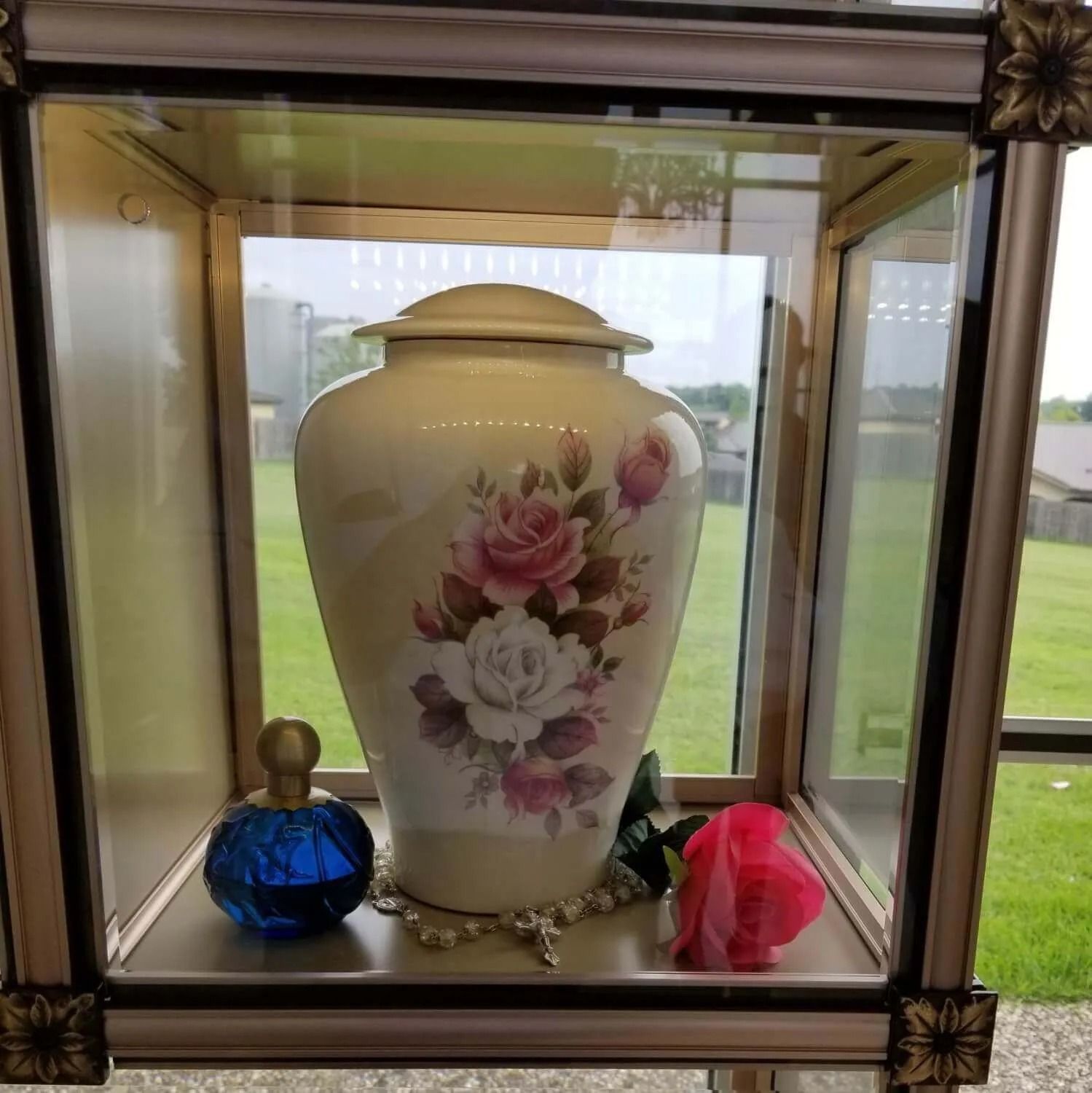 Cream urn with pink roses in a glass display case with decorative elements.