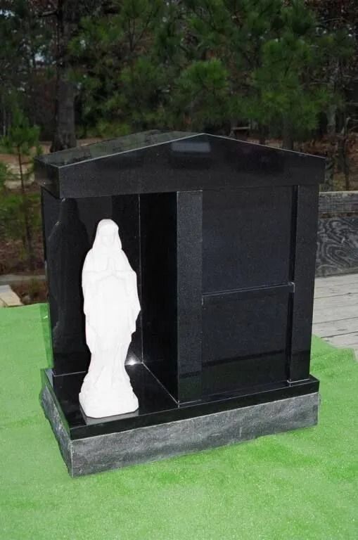 Black granite memorial structure with a white statue of the Virgin Mary inside, set on a stone base, outdoors.