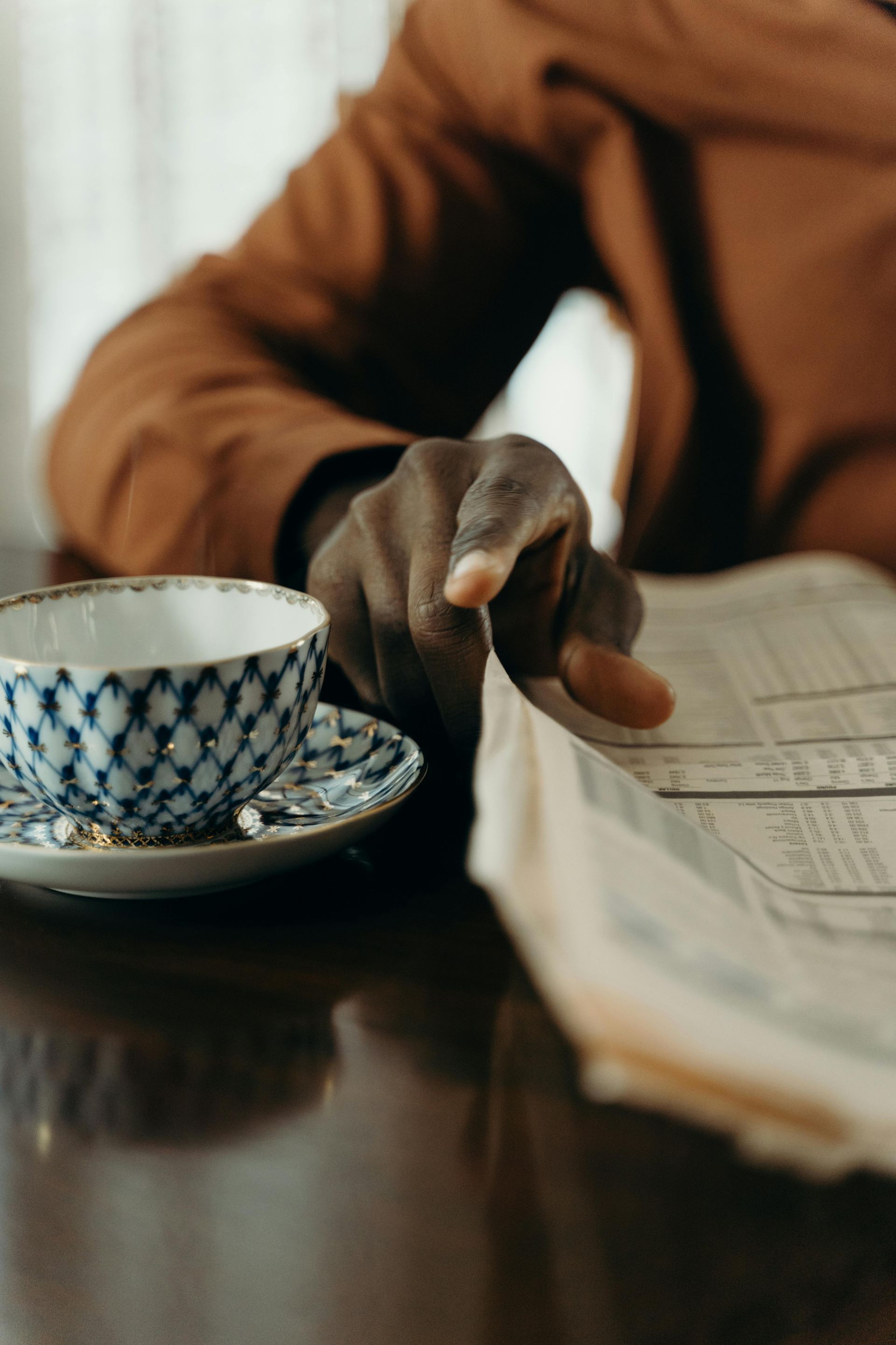 A person's hand pointing at a newspaper. Beside the hand is a teacup on a saucer.