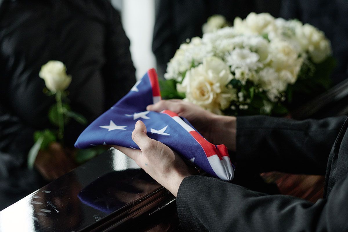 Hands folding a US flag at a funeral, with white flowers in the background.