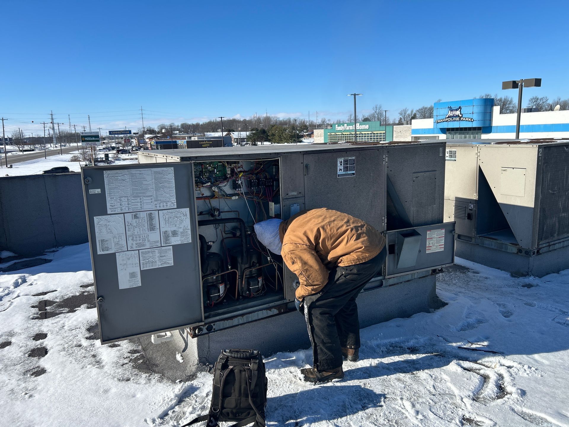 A person in a brown jacket works on the open panel of a rooftop HVAC unit on a sunny, snow-covered roof.