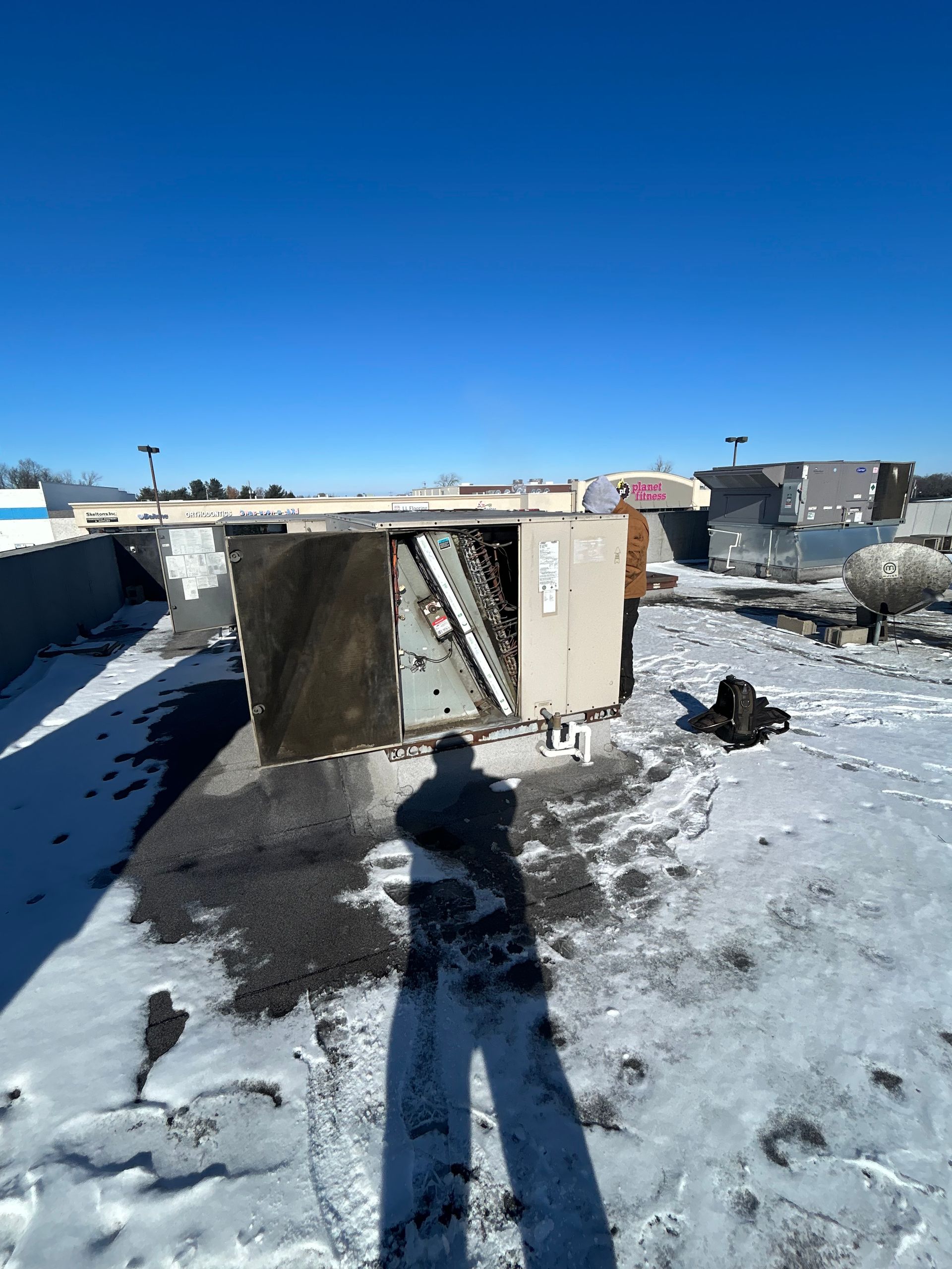 An HVAC unit with an open side panel sits on a flat, snow-covered roof under a bright blue sky.