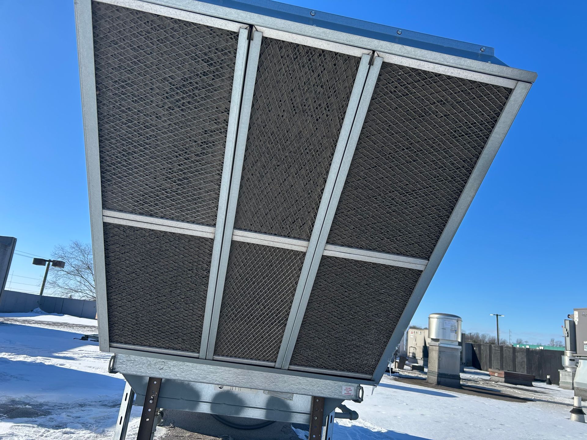 A rooftop cooling unit with large dark honeycomb-patterned air intake panels under a bright blue sky on a snowy day.
