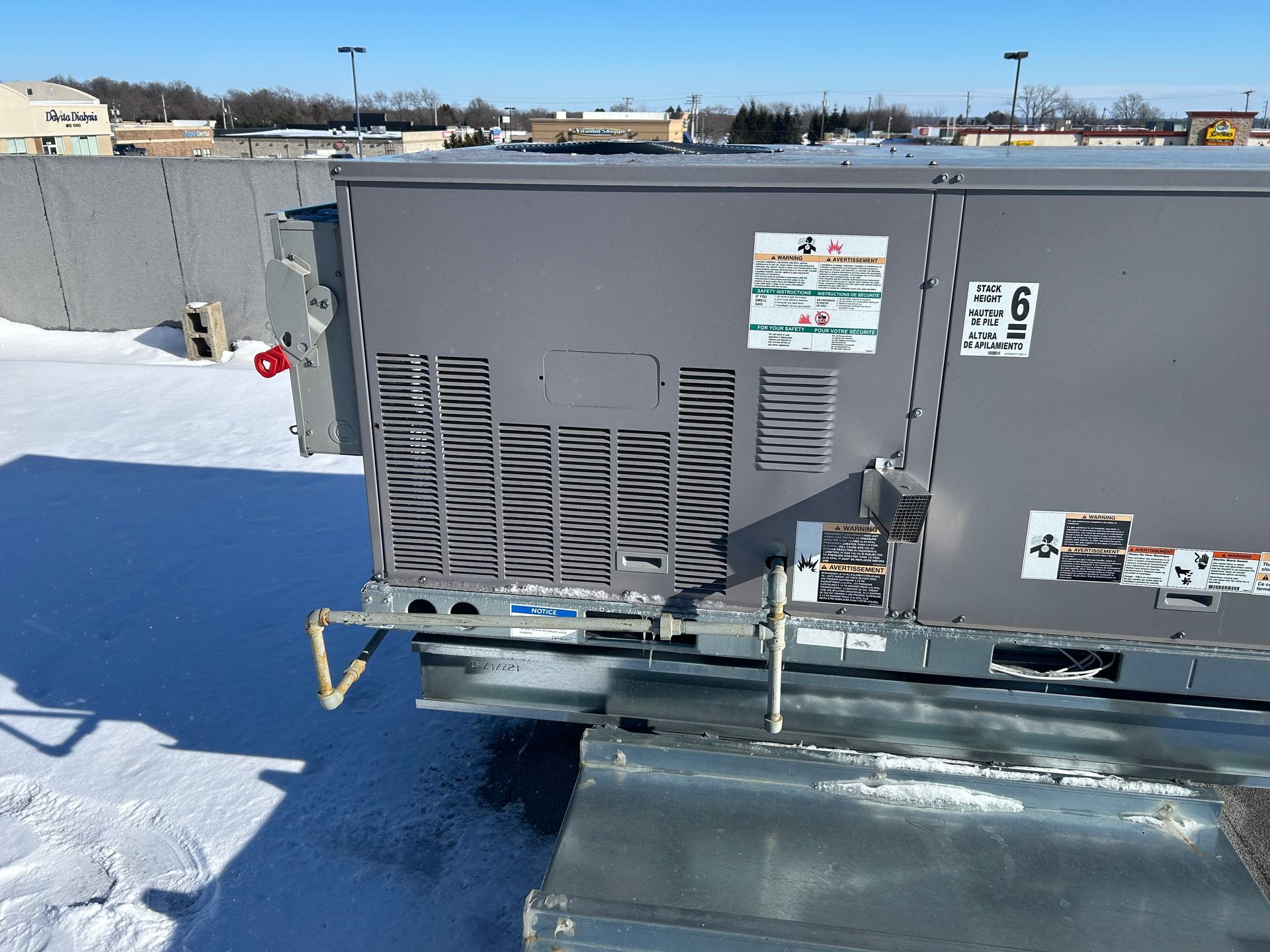 A gray rooftop HVAC unit sits on a snowy roof under a clear blue sky, featuring visible gas piping and maintenance labels.