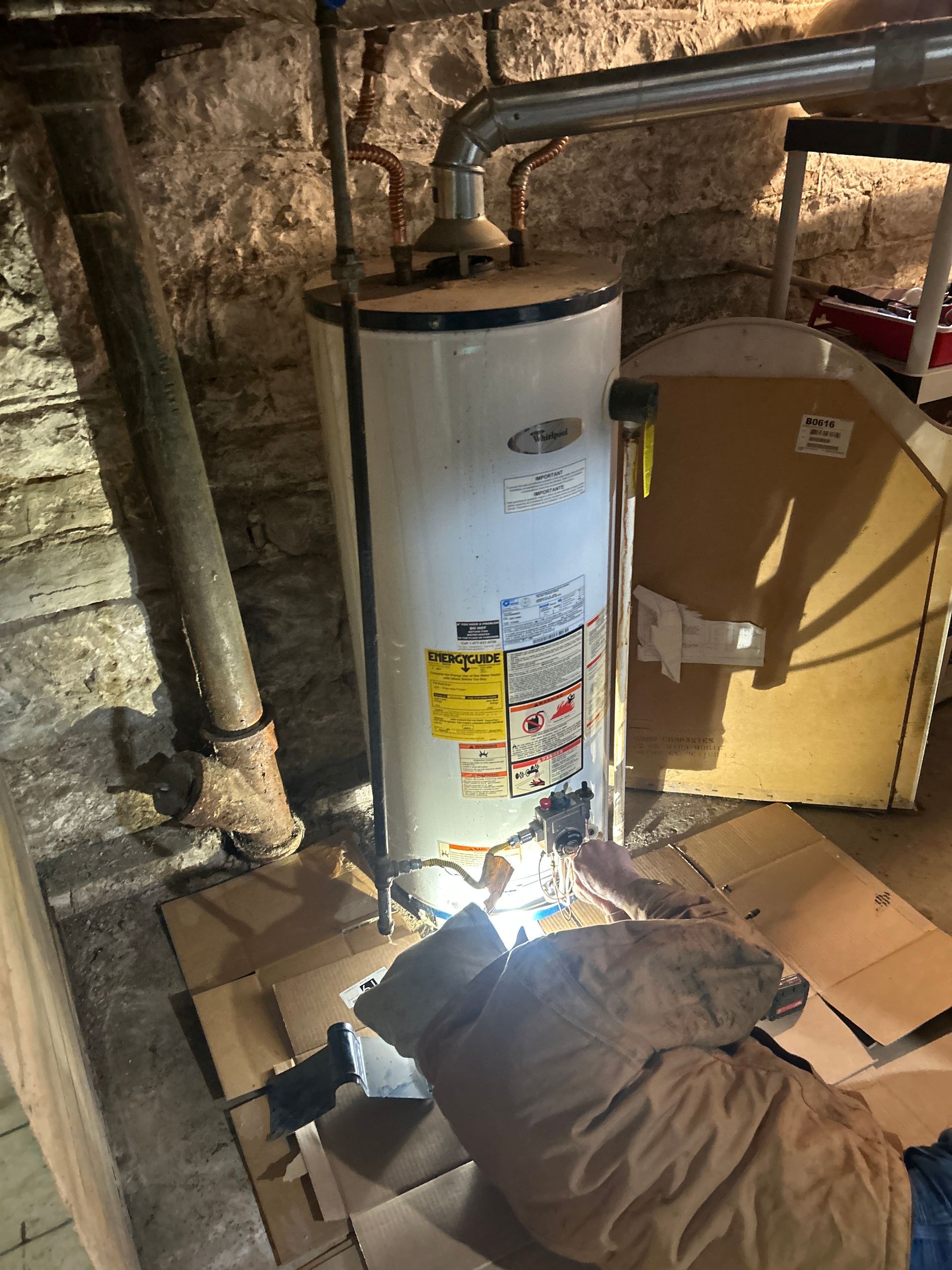 A person works on the base of a white water heater in a basement with stone walls.