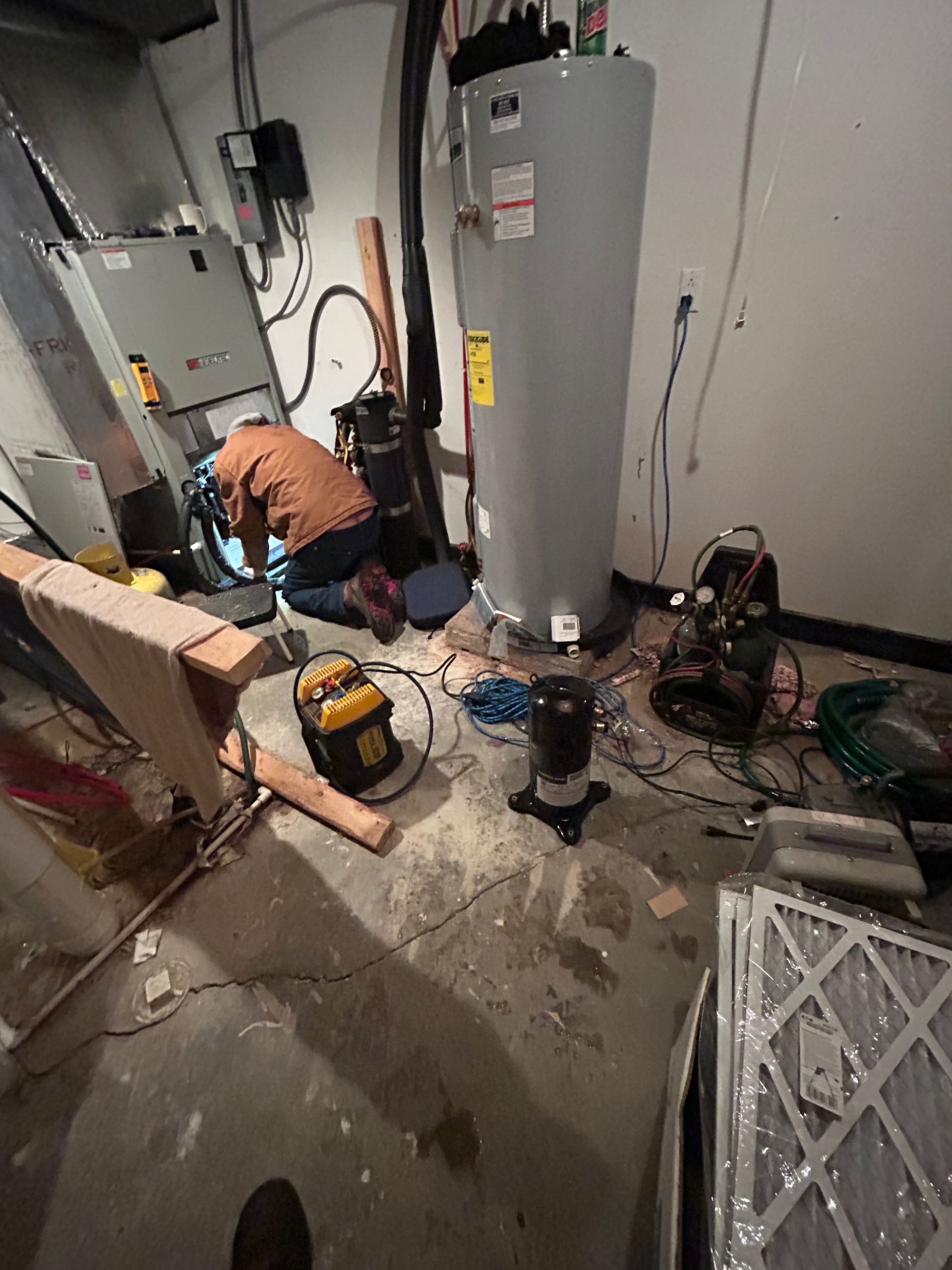 A technician kneels while repairing an HVAC unit in a basement near a water heater and tools.