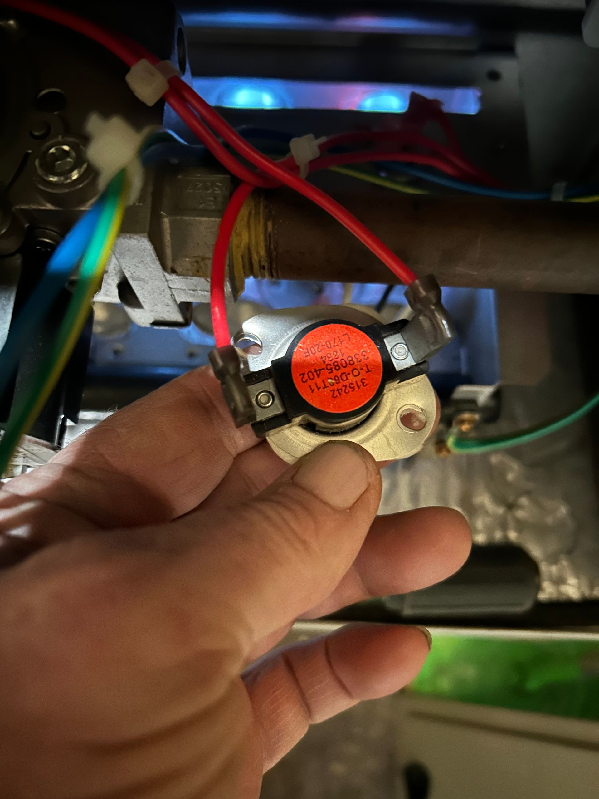 A hand holds a circular thermal fuse switch with a red label, located inside a furnace with glowing blue gas flames.