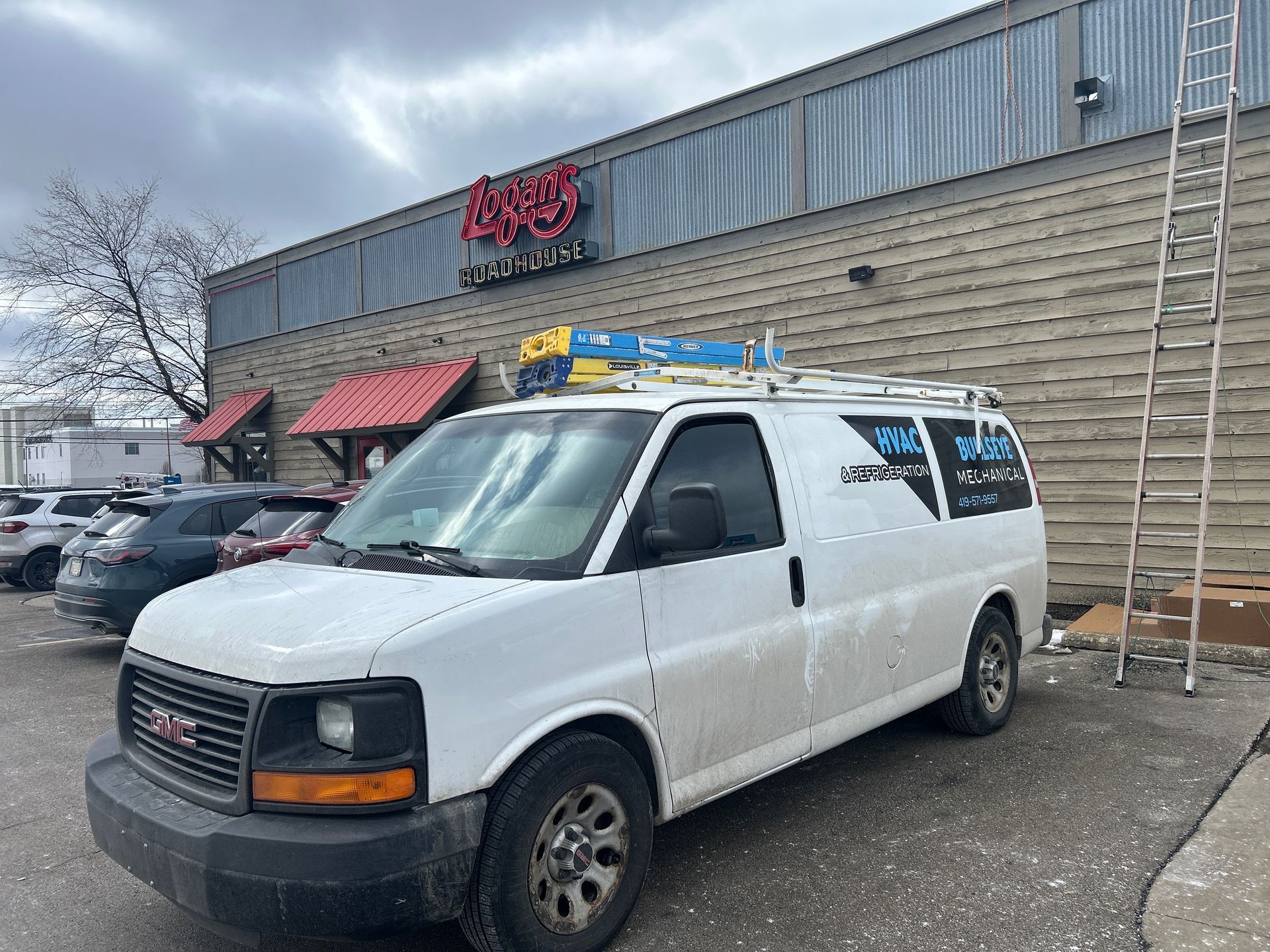 A white work van with a roof ladder rack parked in front of a commercial building with a metal facade and red awnings.