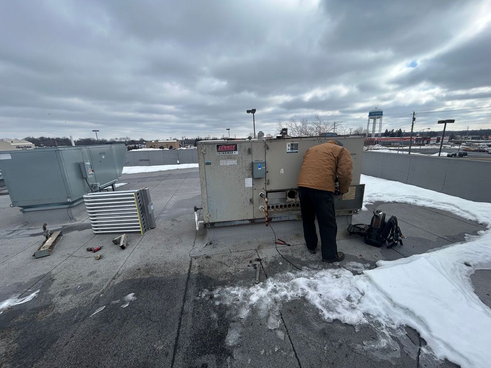 A person in a brown jacket works on a large HVAC unit on a snowy rooftop under a cloudy sky.