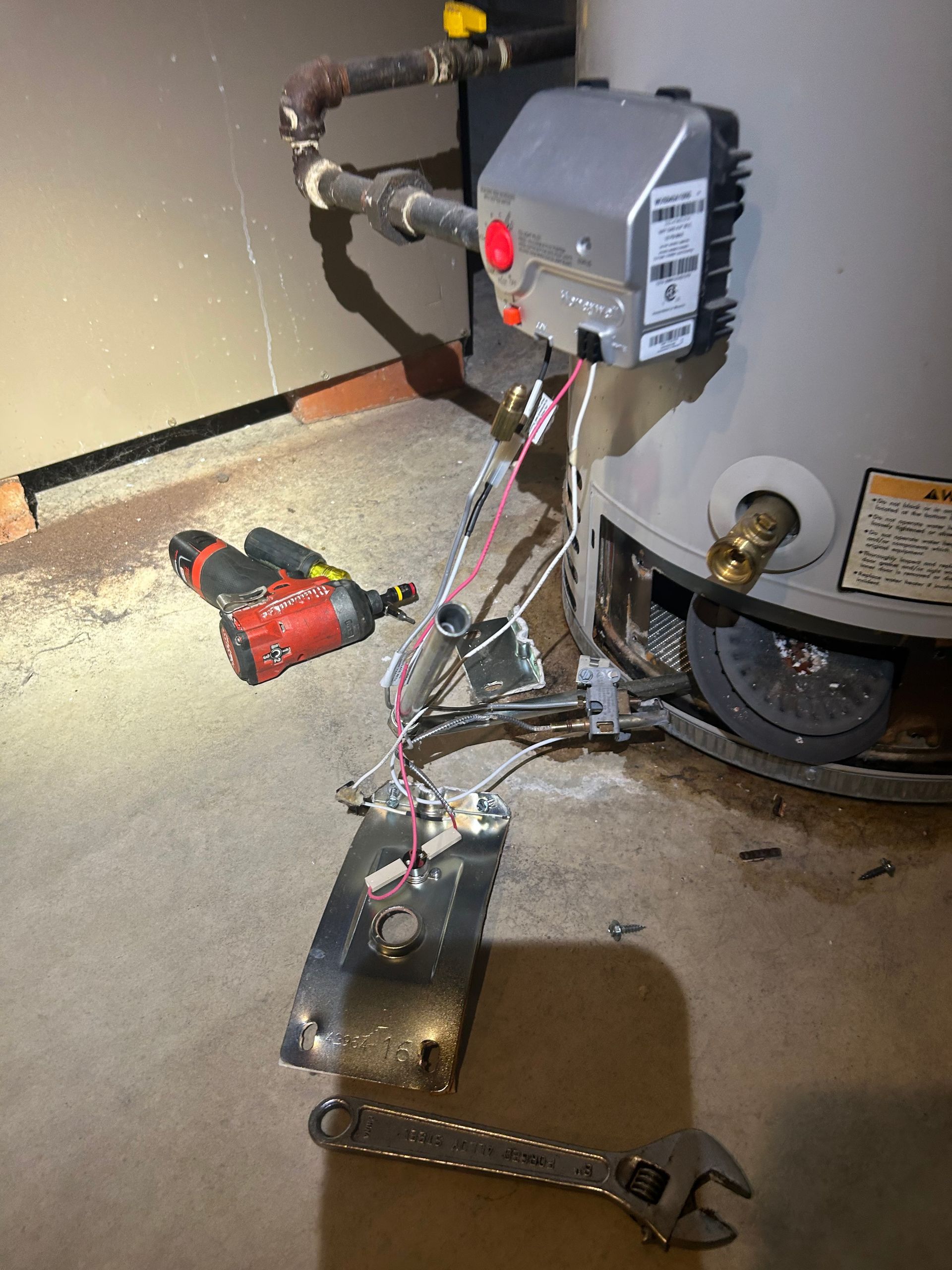 A water heater with its gas control valve and burner assembly removed, sitting on a concrete floor with tools nearby.