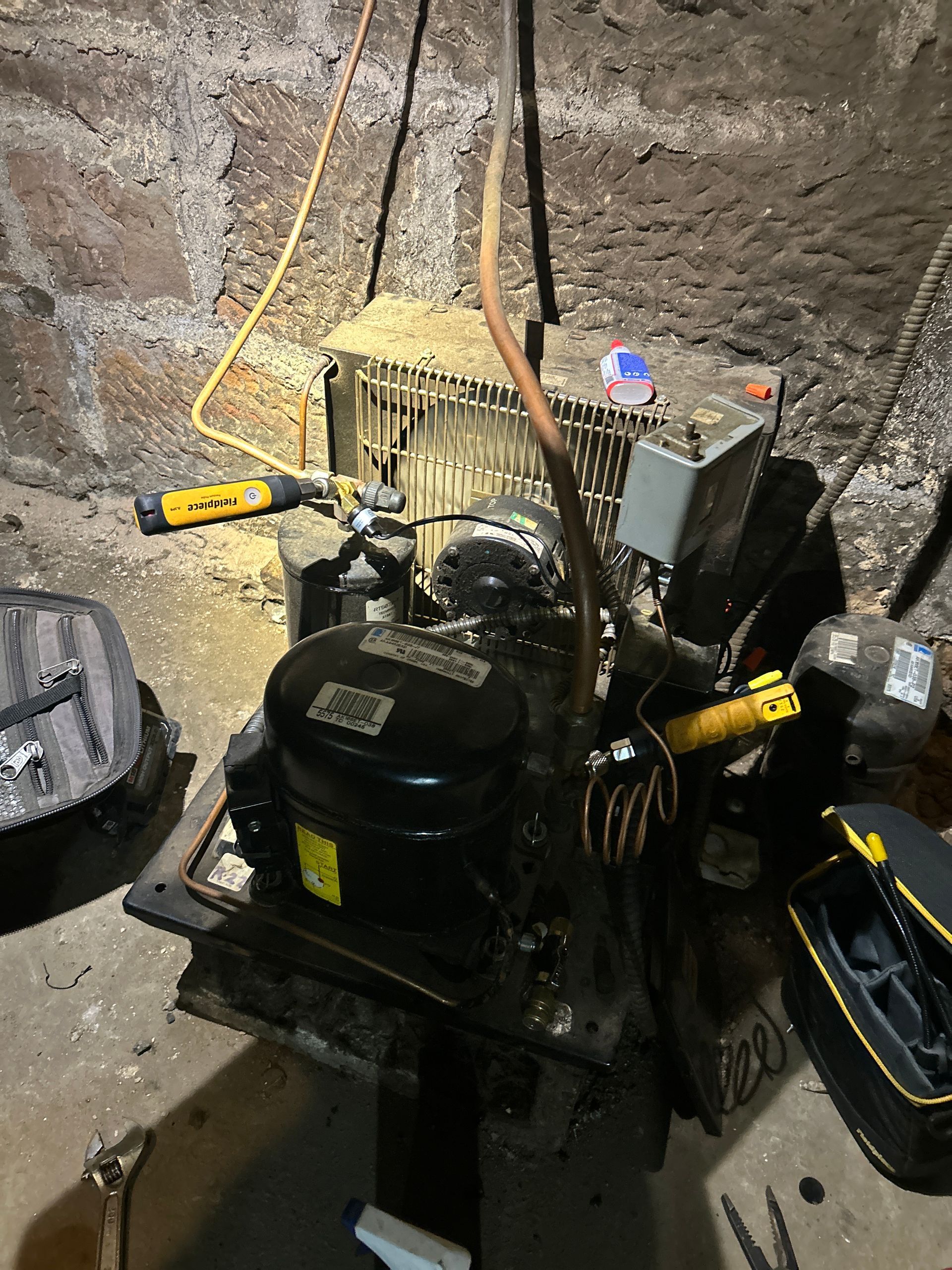 A black refrigeration compressor unit with a metal coil condenser sits on a concrete floor in a basement.