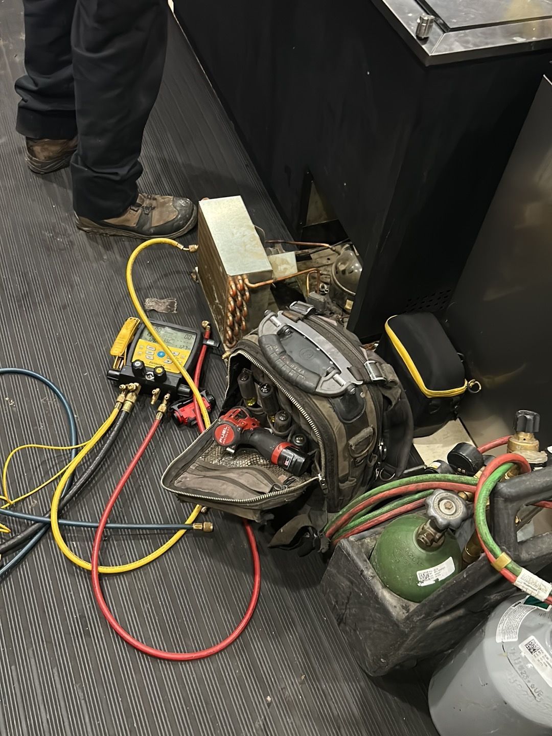 Technician’s tools, including a manifold gauge, tool bag, and nitrogen tank, set on a floor during HVAC repair work.