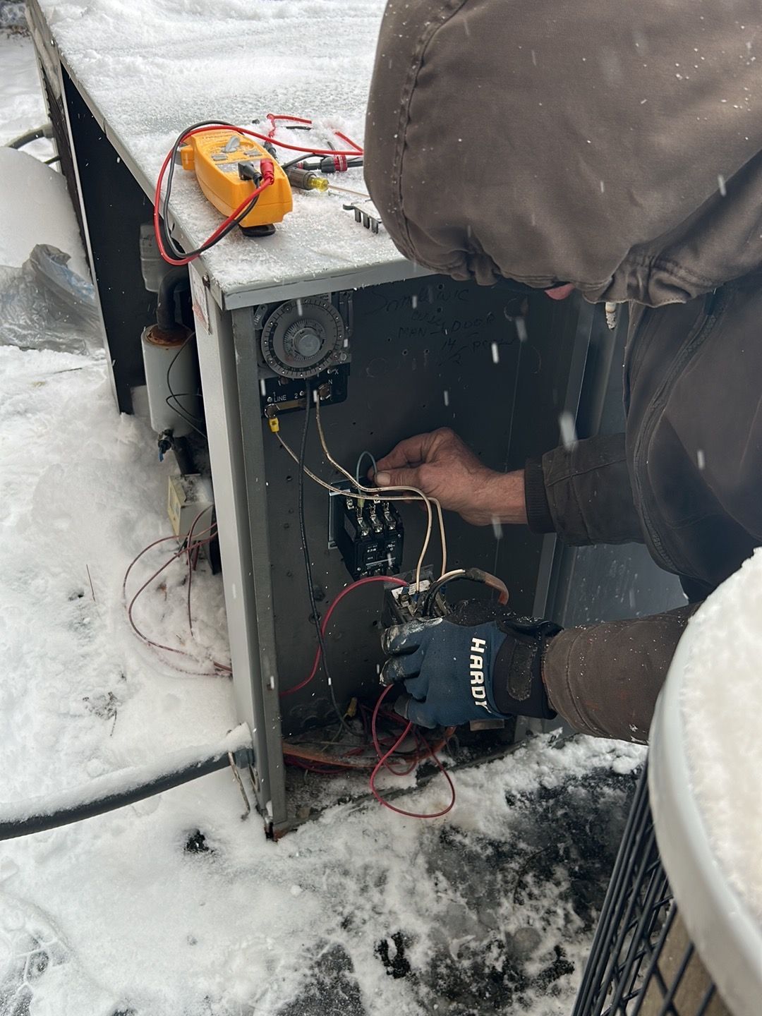 A technician in a hooded jacket repairs an outdoor HVAC unit during a snowstorm, holding wires near a terminal block.