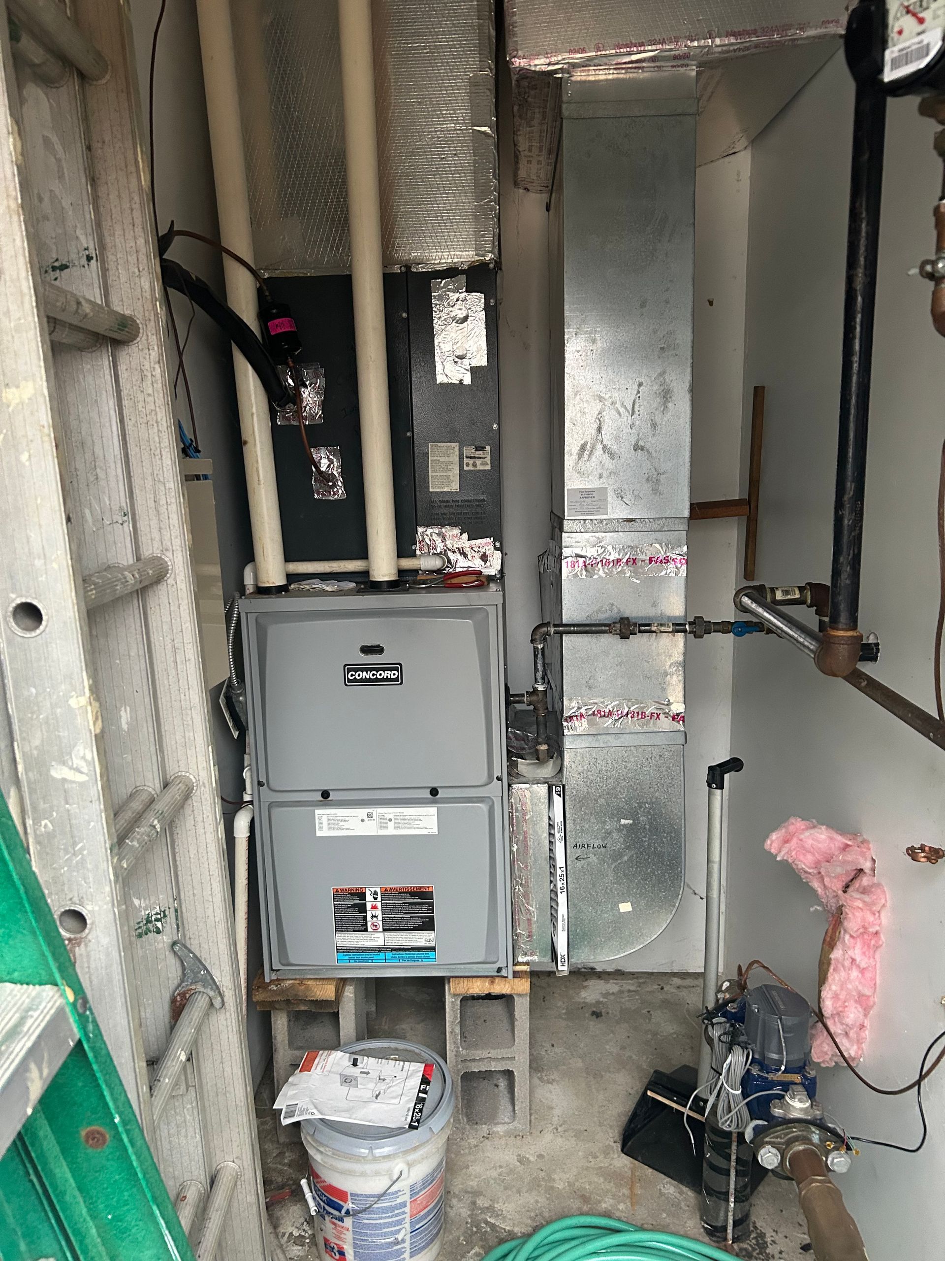 A grey HVAC furnace unit installed in a utility closet, with connected ductwork, pipes, and a bucket placed below.