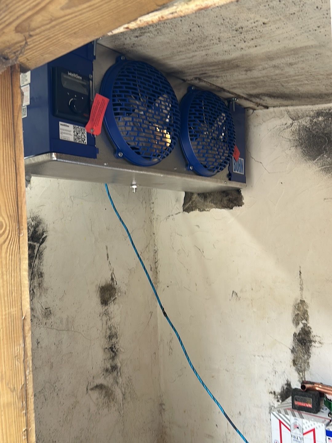 A dual-fan refrigeration unit mounted to the ceiling of a concrete room with a small, blue-cabled control panel.