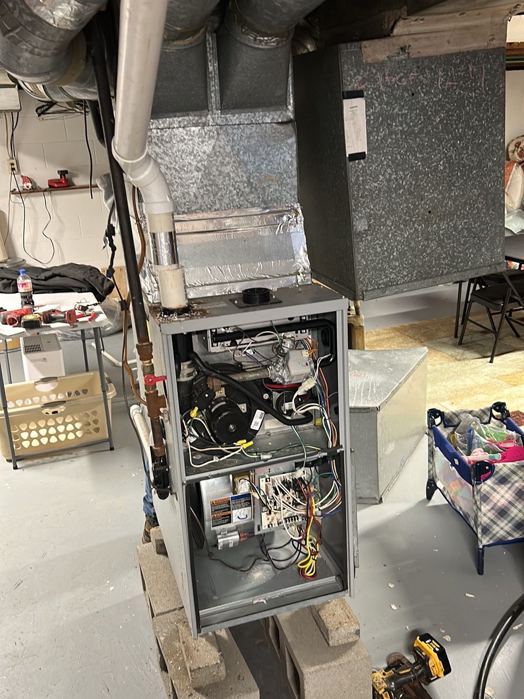 An HVAC furnace with its front panel removed, sitting on cinder blocks in a basement workshop setting.