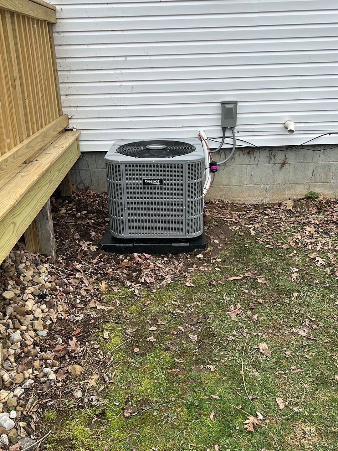An outdoor HVAC condenser unit sits on a black pad beside a white-sided house with a wooden deck.