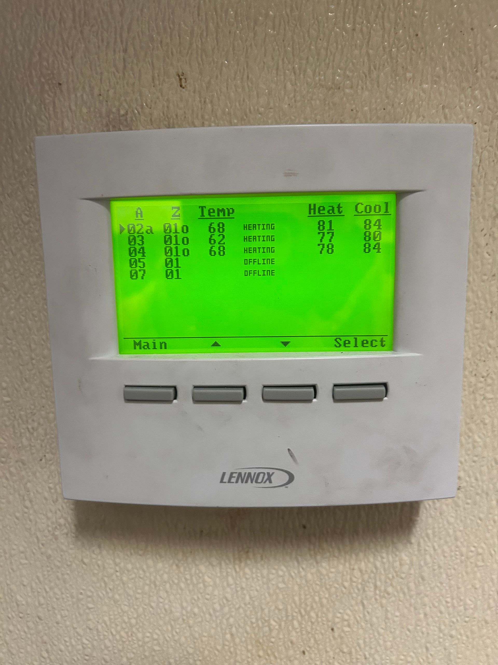 A Lennox digital thermostat mounted on a wall with a backlit green screen displaying zone temperature and settings.