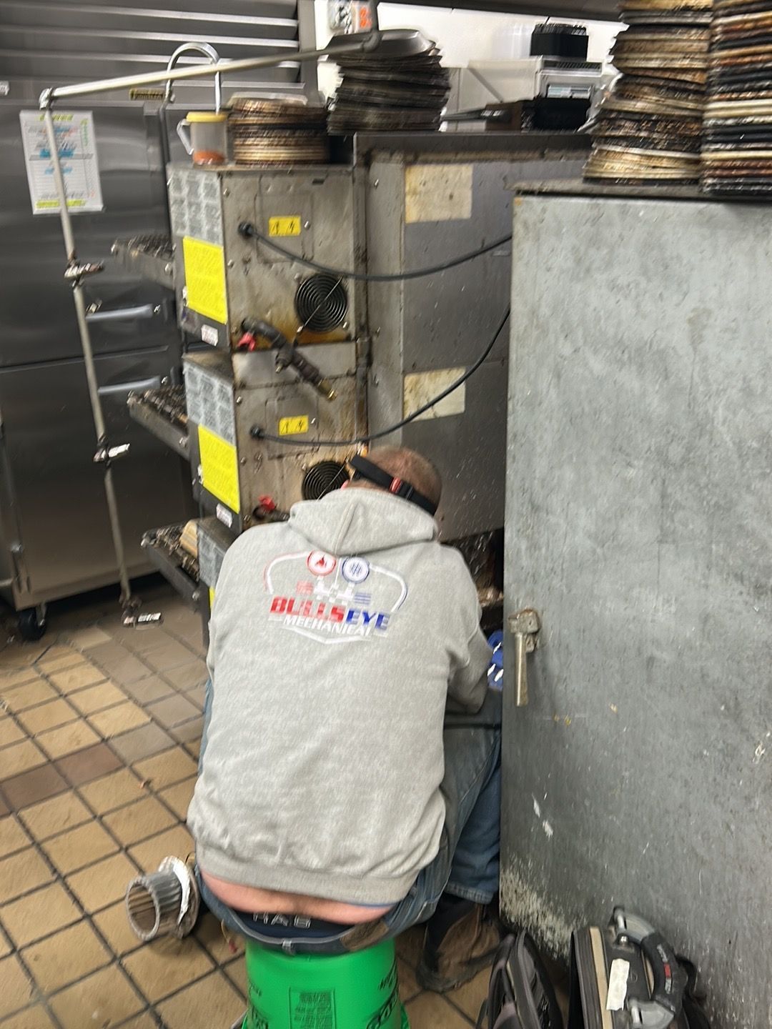A person in a gray hooded sweatshirt sits on a green bucket, working on the back of a large industrial kitchen appliance.